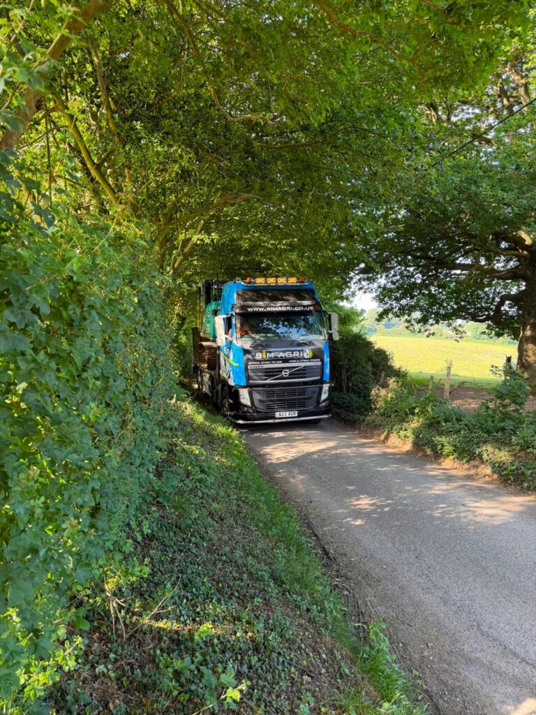 Sims lorry transporting construction plant along a tree-lined road for the plant hire gallery in the UK